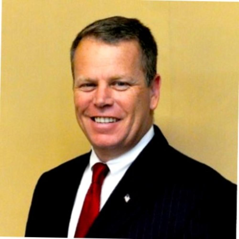 A headshot of Kevin Loncher, who is a man wearing a dark suit with a red tie in front of a yellow background.