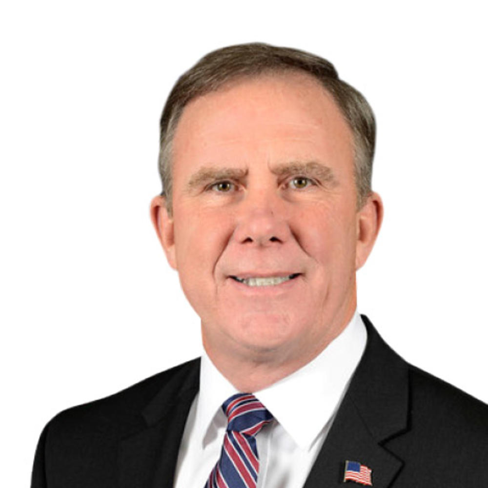 A headshot of Robert McMahon, who is a man wearing a dark suit with a red and blue striped tie in front of a white background.